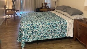 Una camera da letto con una coperta a fiori, un pavimento in legno, una scrivania con una sedia, un comodino con una lampada e un quadro appeso al muro.