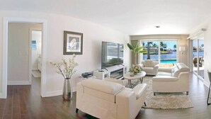 Ein großzügiges Wohnzimmer mit Fernseher, weißen Sofas, einem Couchtisch und Blick auf einen Pool und Palmen.