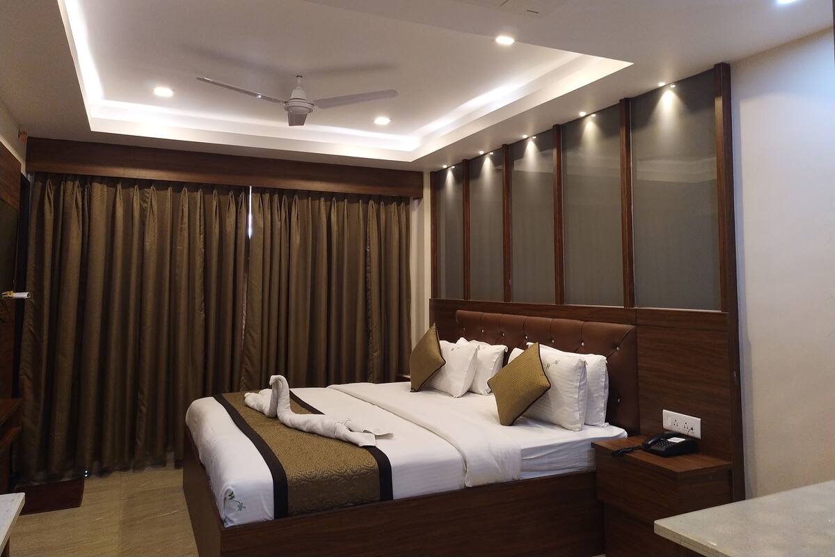 A hotel room with a bed, a bedside table with a phone, a ceiling fan, and a large window with curtains.