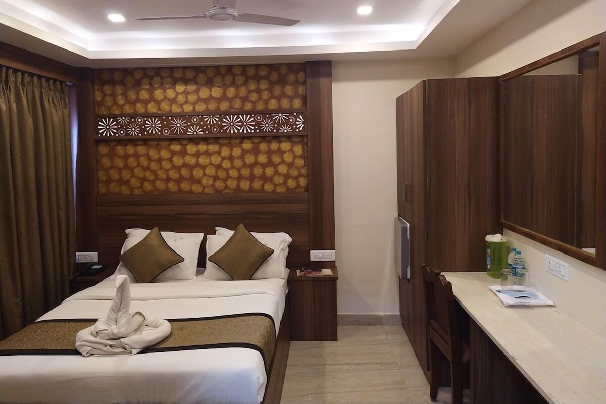 Quarto de hotel com uma cama, escrivaninha, espelho, ventilador de teto e um painel decorativo na parede.