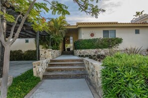 A house with a stone staircase leading to a door, surrounded by greenery and a pergola.