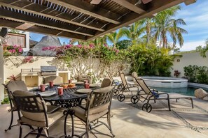 An outdoor patio with a dining table, chairs, and a grill. There is a pool and palm trees in the background.