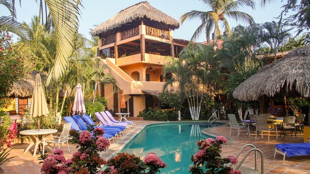 A pool area with lounge chairs, a thatched-roof building, and tropical plants. - Lg 1 BR apt w/ ionization system pool, free yoga classes, gardens, spa service (Puerto Escondido)