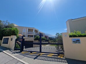 Une entrée fermée par un portail mène à un immeuble, le ciel est dégagé et le soleil brille.