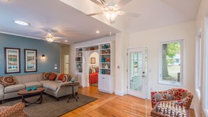 A spacious living room with a sectional sofa, a coffee table, and a chess table. There are ceiling fans, a bookshelf, and a door leading to a patio.