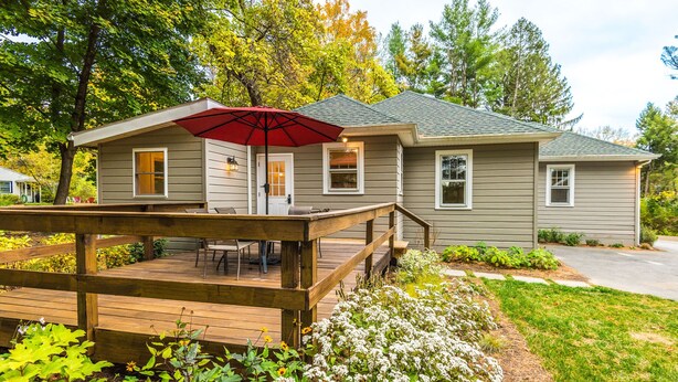 A two-story house with a deck, a patio with a red umbrella, and a front yard with various plants.