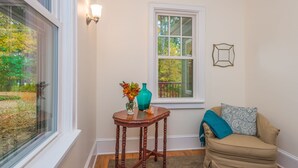 A corner room with a window, a small table with a vase and a blue vase, a beige armchair with a patterned pillow, and a wall-mounted light fixture.