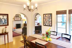 A dining area with a round table, chairs, and a chandelier. There are framed pictures on the walls and a window with blinds. - BEACH BUNGALOW IN THE HEART OF SURF CITY 5BR 3BA - Work place friendly (Huntington Beach)