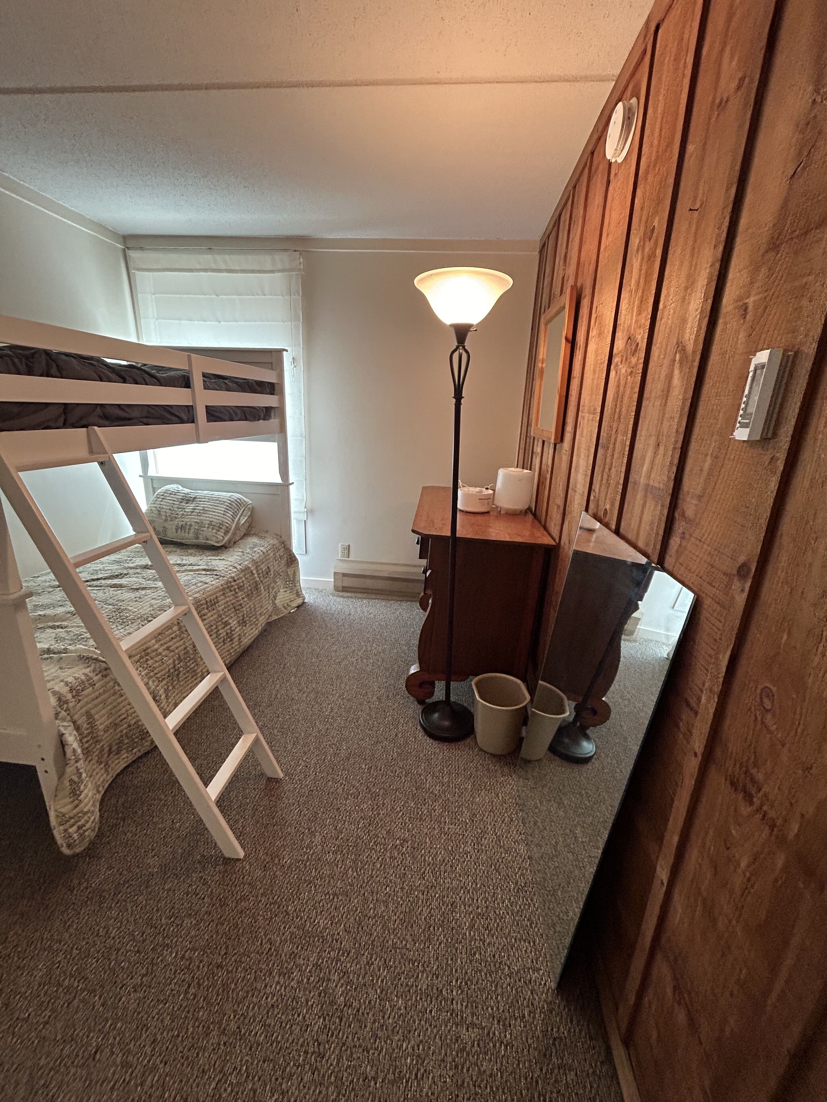 A bunk bed room with a ladder, a desk with a lamp, and a wooden wardrobe.