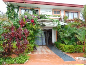 Enter a serene oasis with a checkered pathway leading to a glass door, surrounded by lush greenery and vibrant plants.