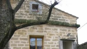Ein Steinhaus mit einem Fenster und einem Rundbogenportal. Vor dem Gebäude stehen Pflanzgefäße mit Pflanzen.