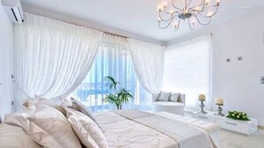 A modern bedroom with a large bed, white curtains, a chandelier, and a view of the outdoors. - Luxury White  Suite(2 Persons) (CHANIA)