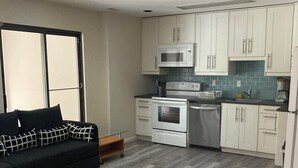 A compact kitchen with white cabinets, a microwave, stove, and a dishwasher. There is a black sofa with patterned cushions and a small wooden side table.