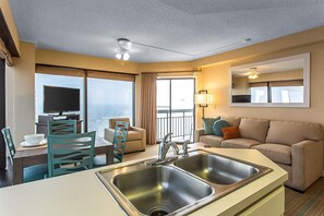 Une chambre d’hôtel moderne dotée d’une kitchenette, d’un canapé, d’une table à manger et d’une grande fenêtre donnant sur l’océan.