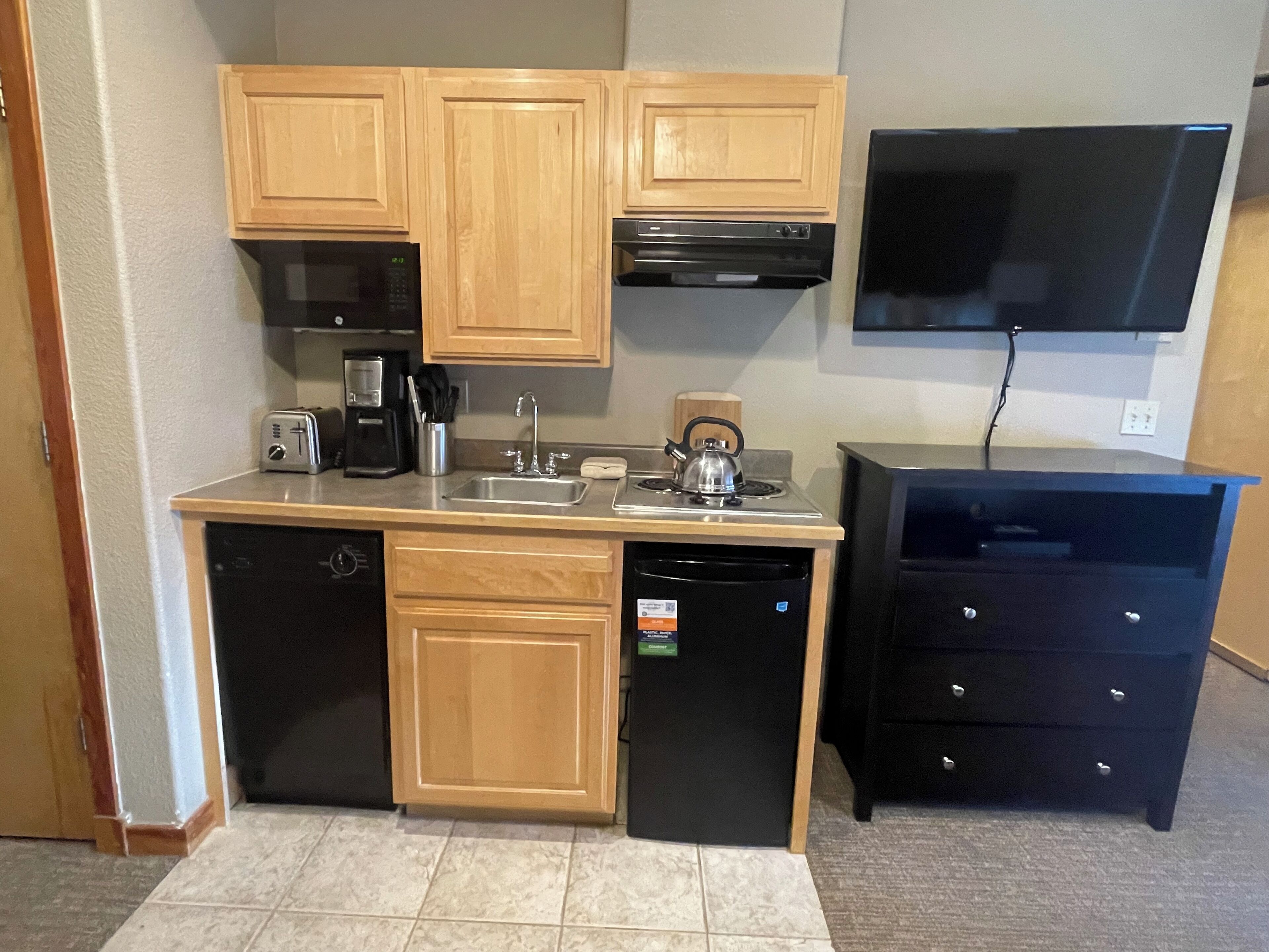A compact kitchen with wooden cabinets, a dishwasher, a coffee maker, a kettle, and a black refrigerator.