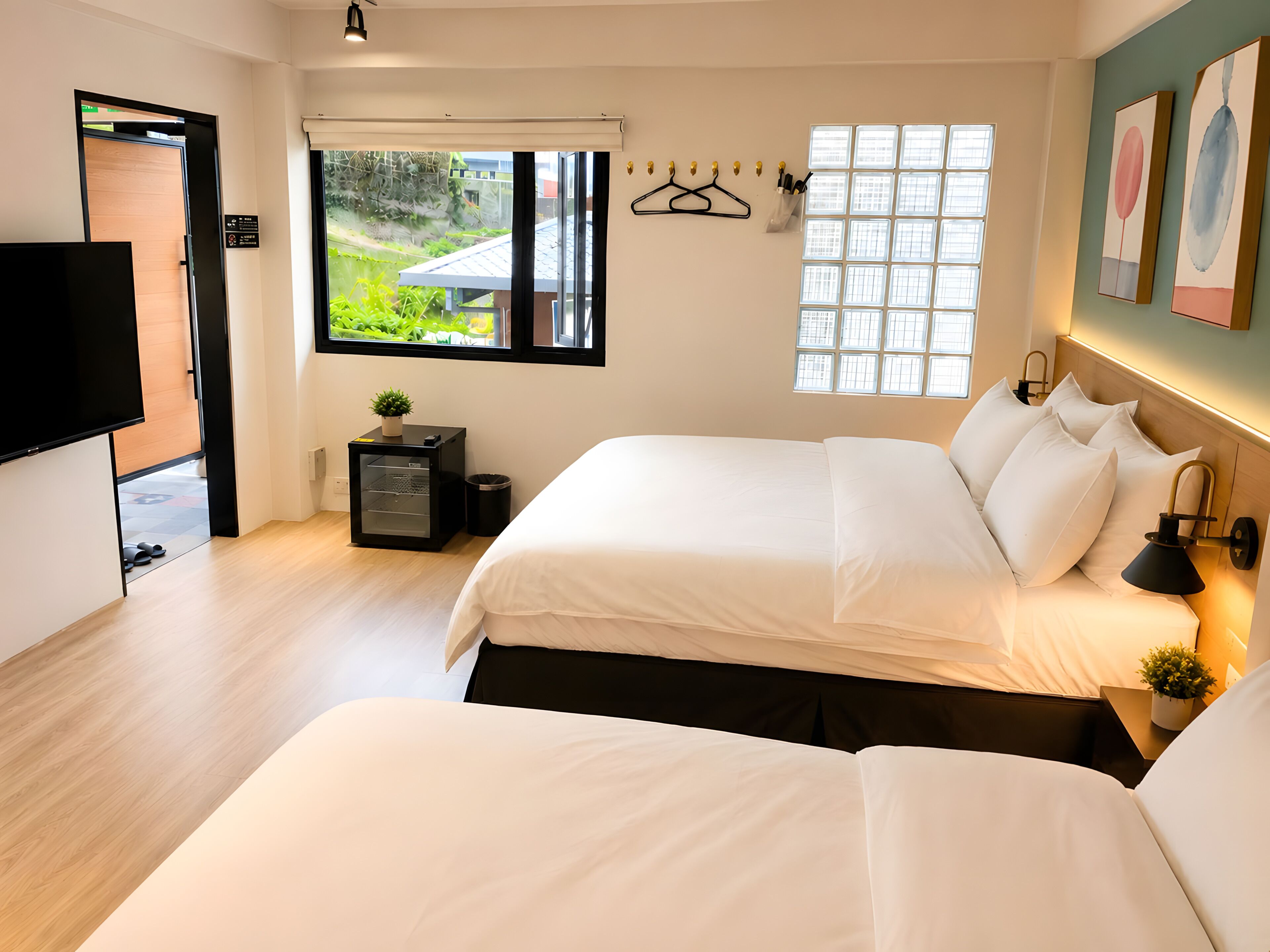 A modern hotel room with two beds, a TV, a small desk, and a window with a view of greenery.