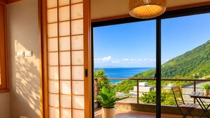 Ein Zimmer mit einer verschiebbaren Shoji-Schirmwand, einem Bett, einer Topfpflanze und einem Balkon mit Blick auf das Meer und die Hügel.