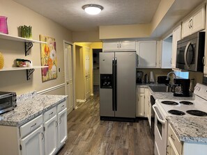 A modern kitchen with white cabinets, a stainless steel refrigerator, a microwave, a stove, and a countertop with a granite-like surface.