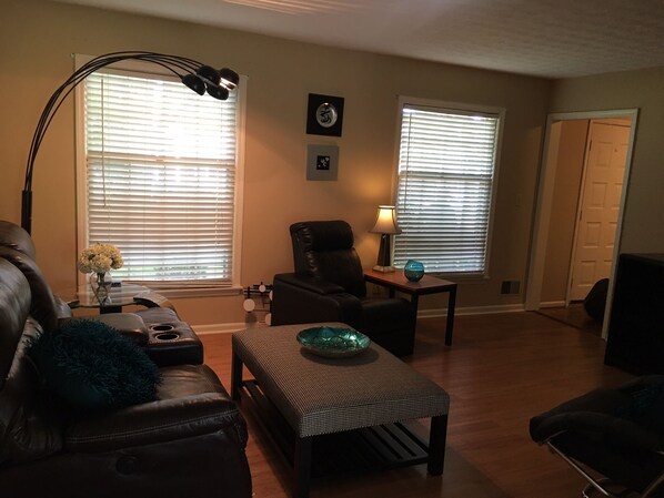 A living room with a leather sofa, a patterned ottoman, a glass coffee table, a black armchair, and a floor lamp.