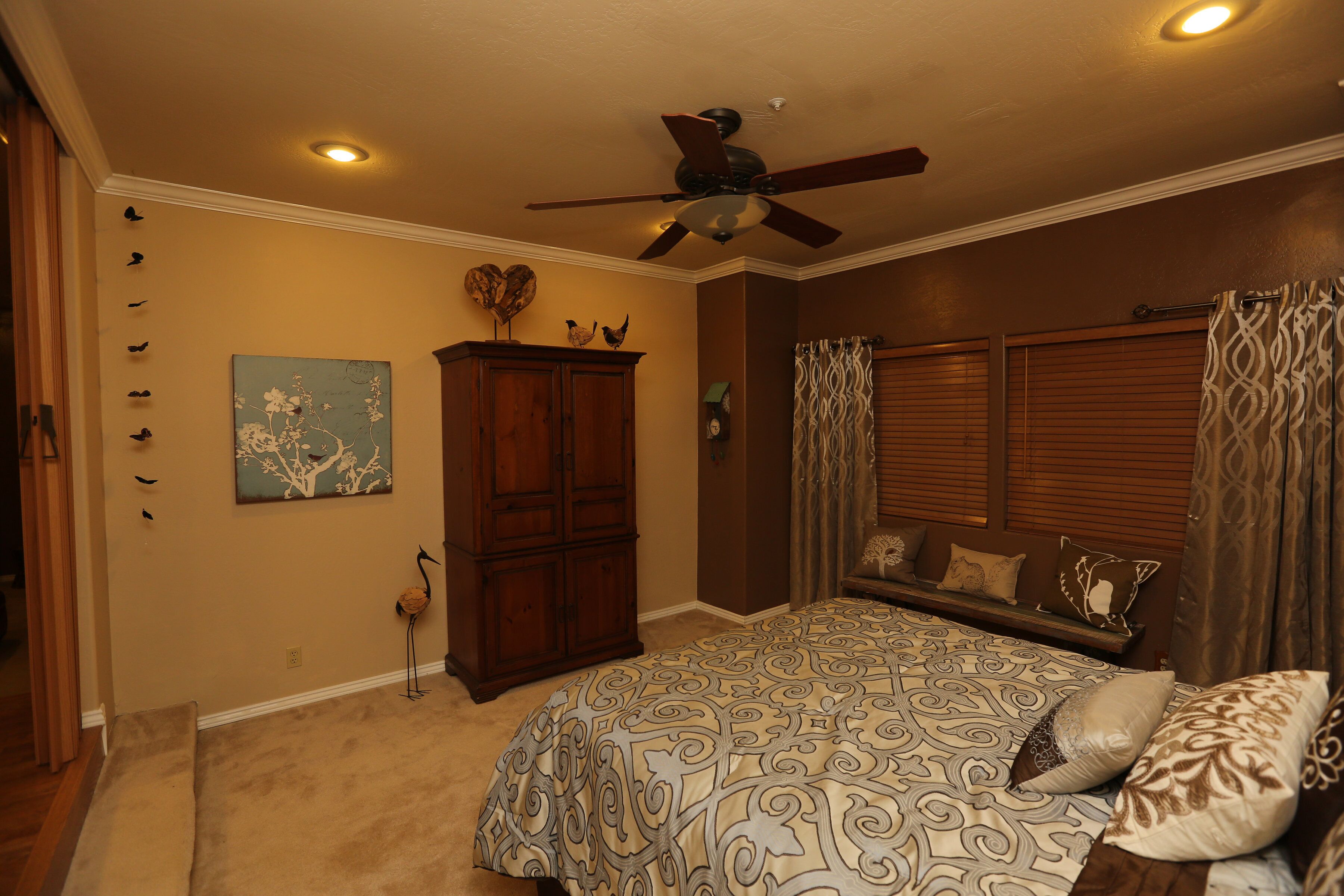 A bedroom with a large bed, a ceiling fan, a wooden wardrobe, and a wall-mounted clock.