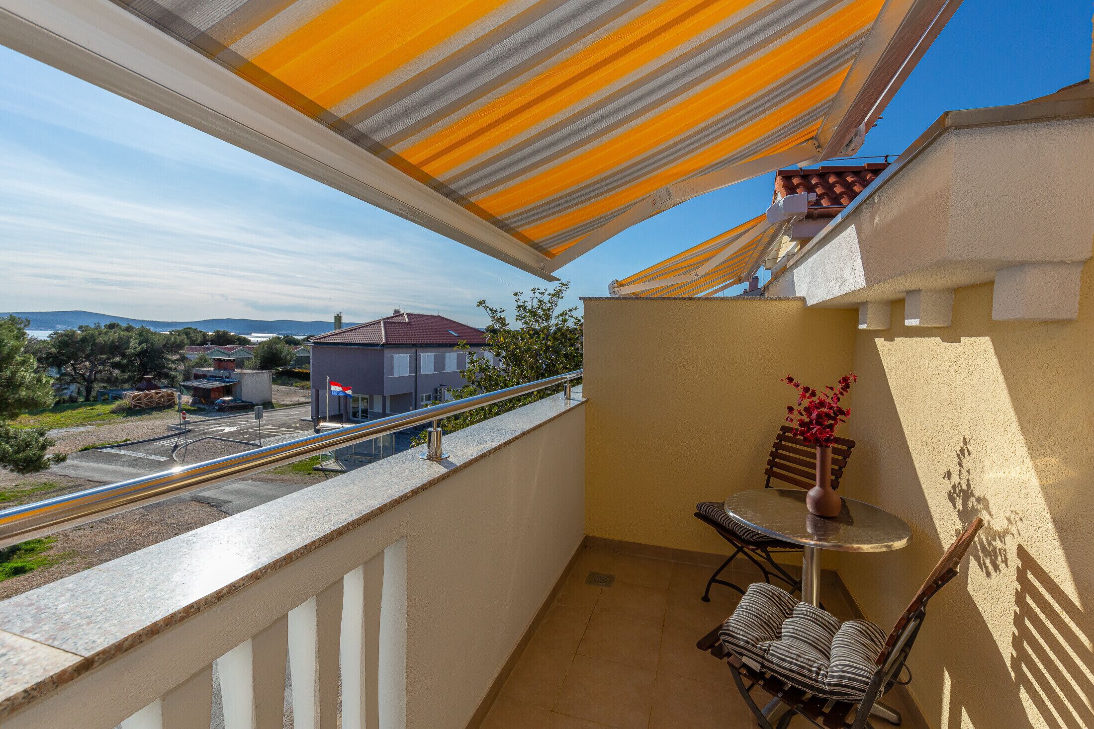 A balcony with a view of a road and buildings, a table with chairs, and a vase with flowers.