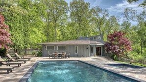 Una piscina con lettini e zona pranzo, immersa tra alberi e una casa in legno.