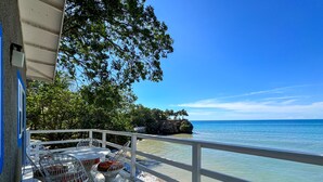A wooden deck with white railings overlooking a clear blue sea, featuring wicker chairs and a table.