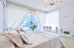 A modern bedroom with a large bed, white curtains, a chandelier, and a view of the outdoors. - ktima reveli luxury villa (χανια καλαθας)