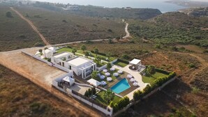 An aerial view of a modern resort with a swimming pool, outdoor seating, and a scenic hillside backdrop. - ktima reveli luxury villa (χανια καλαθας)
