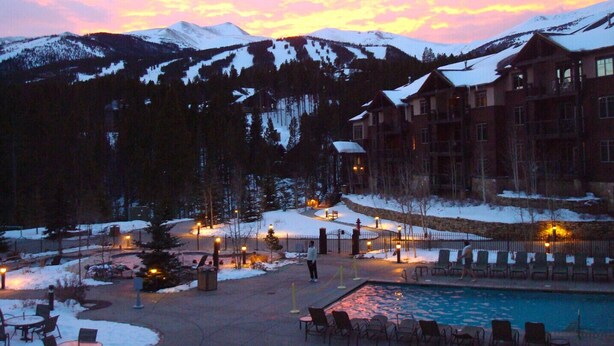 A resort with a pool, outdoor seating, and a view of snow-covered mountains at dusk.