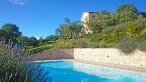 Une piscine aux murs en pierre, entourée de plantes de lavande et bordée d’une colline sur laquelle se trouve un bâtiment en arrière-plan.