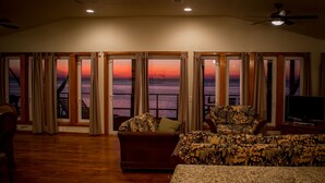 Un salon avec vue sur l’océan au coucher du soleil, un plancher en bois et un téléviseur posé sur un meuble.