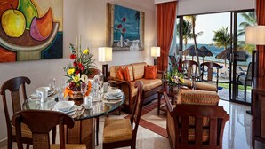 Un coin repas avec une table ronde dressée pour six personnes, entourée de chaises en bois. Un canapé et des fauteuils sont placés au premier plan, offrant une vue sur l’océan et les palmiers à travers de grandes fenêtres.