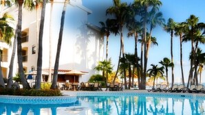 Un espace aménagé au bord de la piscine, avec des chaises longues, des palmiers et un bâtiment doté de balcons.