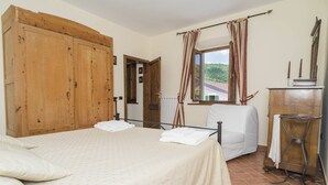 Un dormitorio con cama, armario de madera, una silla, una mesita y una ventana con cortinas.