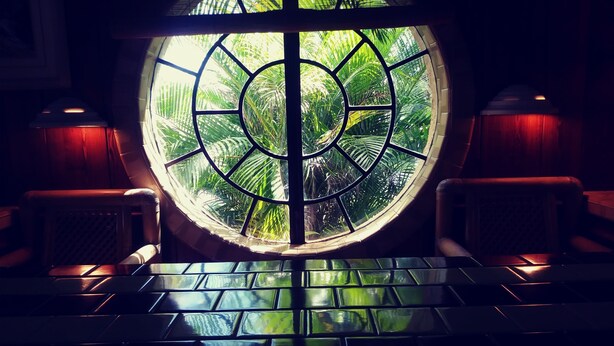 A circular window with a tropical view, a tiled floor, and wooden furniture.