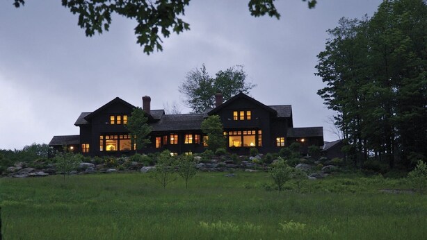 Una casa grande de color oscuro con numerosas ventanas y una chimenea, rodeada de un campo cubierto de césped y árboles.