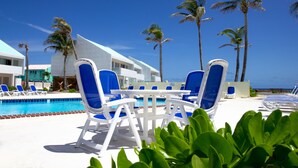 A poolside area with blue and white lounge chairs, a pool, palm trees, and a modern building in the background.