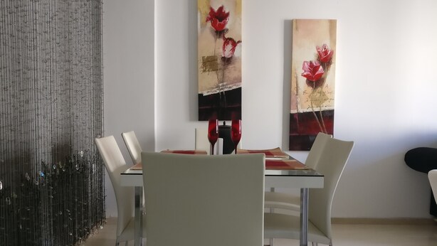 A modern dining area with a glass table, white chairs, and two paintings of red flowers on the wall.