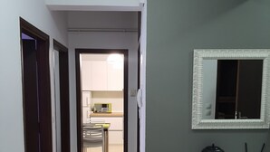 A hallway leading to a kitchen with a microwave and cabinets. There is a desk with a mirror and a small table with a teapot and a cup.