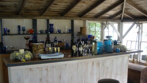 A rustic bar area with wooden beams, shelves with various bottles and glasses, and two stools. - Charming 18th century cottage with its own beachfront on Scargo Lake near bay (Dennis)