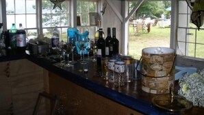 A rustic bar area with wooden beams, a counter with various bottles and glasses, and a view of the outdoors. - Charming 18th century cottage with its own beachfront on Scargo Lake near bay (Dennis)