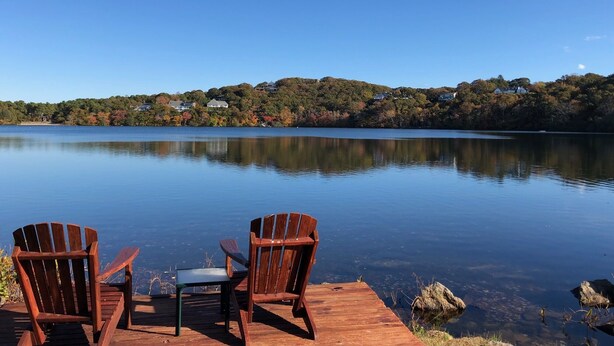 A wooden dock with two Adirondack chairs and a small table overlooking a calm lake with a forested hillside in the background. - Charming 18th century cottage with its own beachfront on Scargo Lake near bay (Dennis)