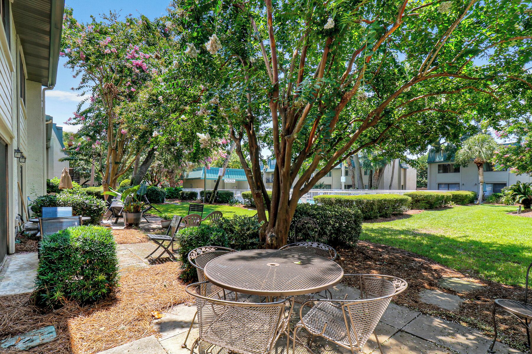 A patio with a metal table and chairs, surrounded by greenery and a tree.
