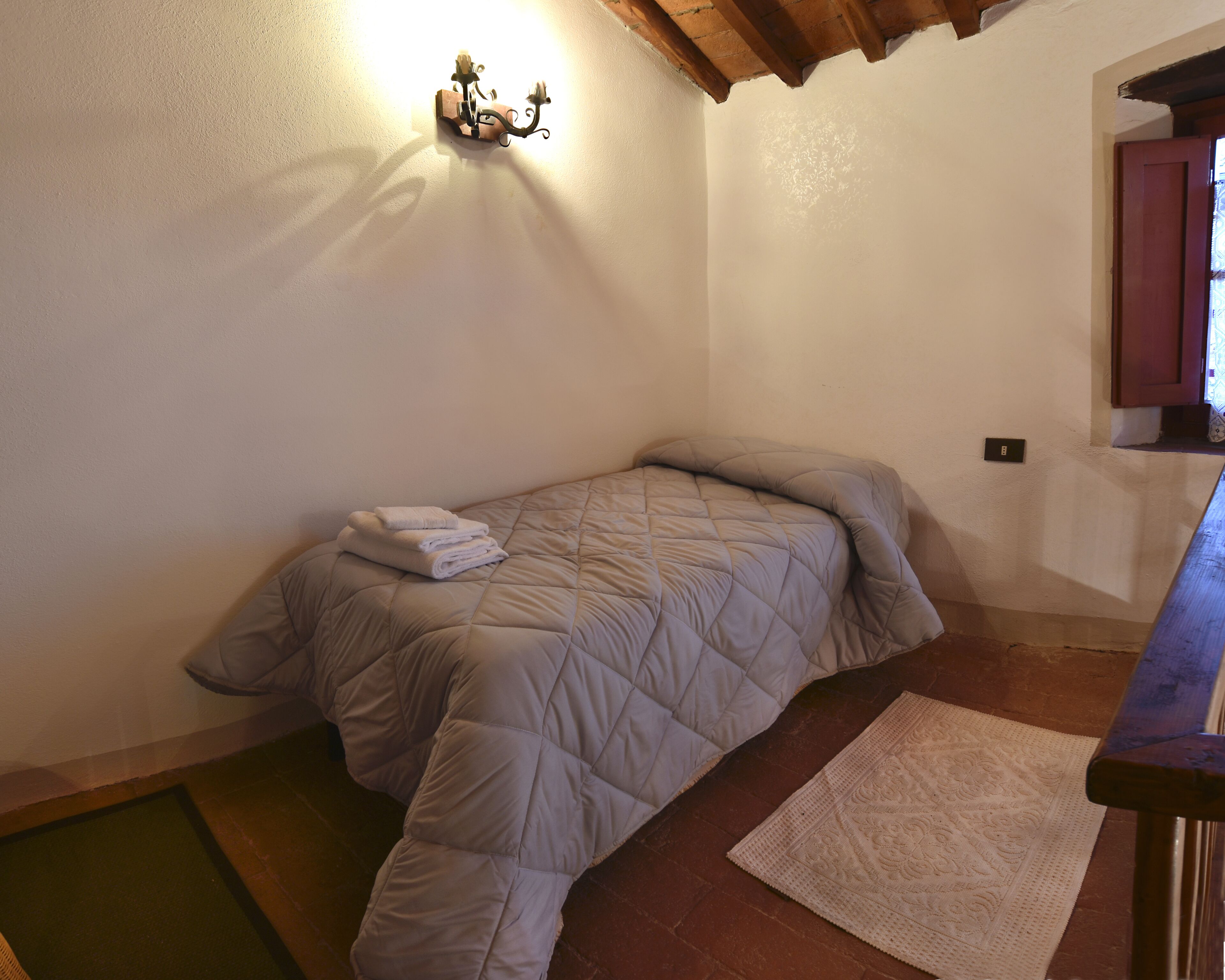 Ein Einzelbett mit gesteppter Decke und gefalteten Handtüchern, eine Holzbank, ein gemustertes Teppich und eine Wandleuchte.