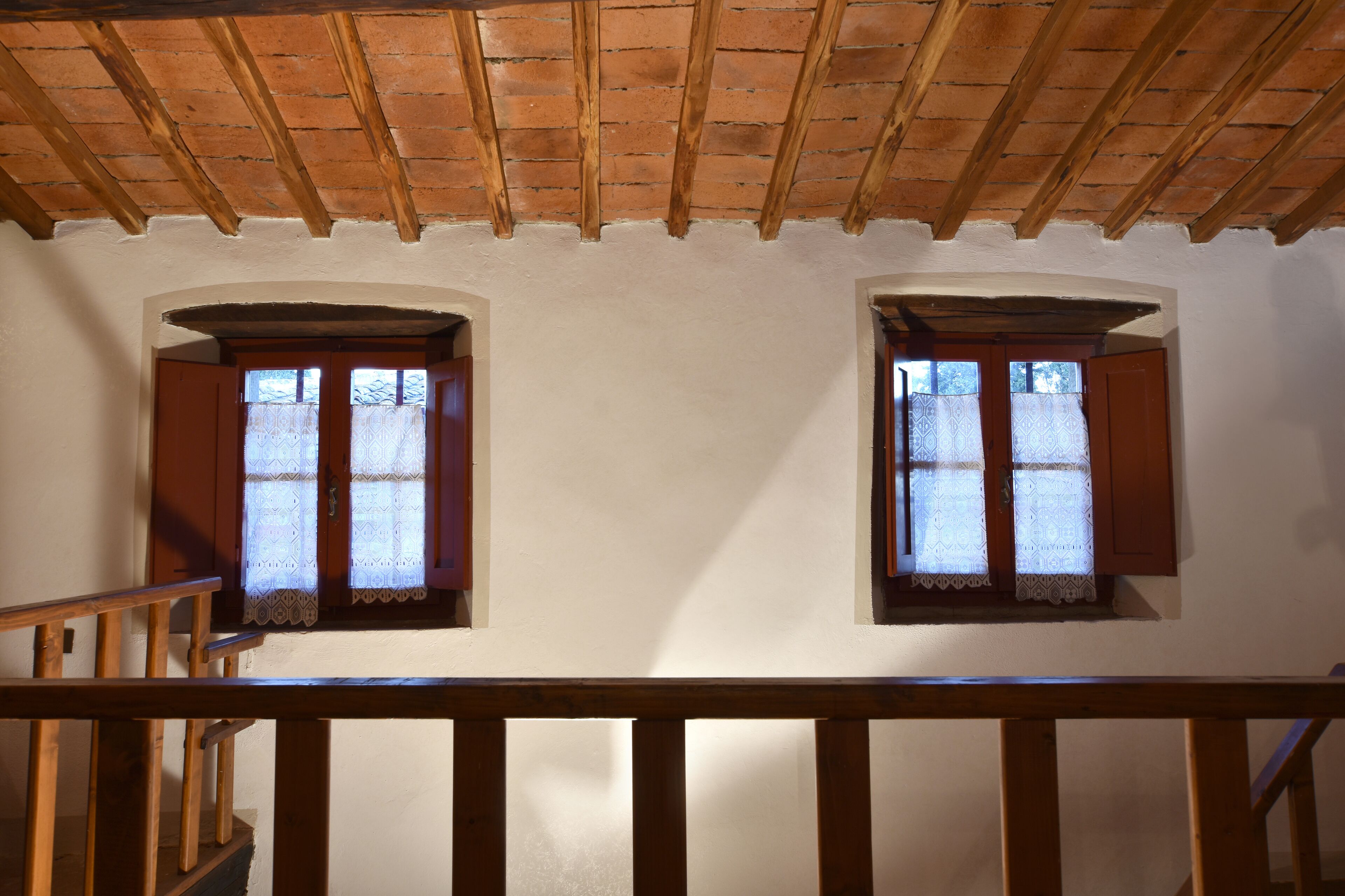 Una habitación con dos ventanas, vigas de madera en el techo y barandilla de madera.