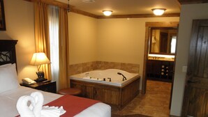 A hotel room with a large bed, a wooden bathtub, a nightstand with a lamp, and a bathroom visible through an open door.