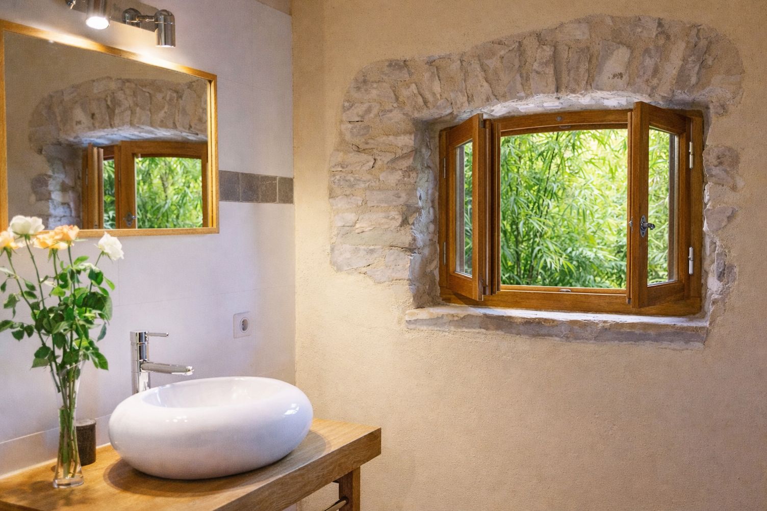 Ein Badezimmer mit Steinwand, Holzfenster, Waschbecken in Weiß und einem Spiegel.