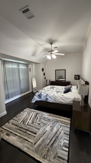 Une chambre à coucher comprenant un lit, un ventilateur de plafond, une lampe de chevet, une commode, un miroir et une fenêtre avec des rideaux.