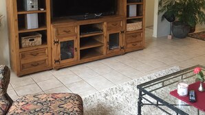 Sala de estar com uma grande TV de tela plana em um móvel de madeira, sofá estampado e mesa de centro com um vaso e controle remoto.